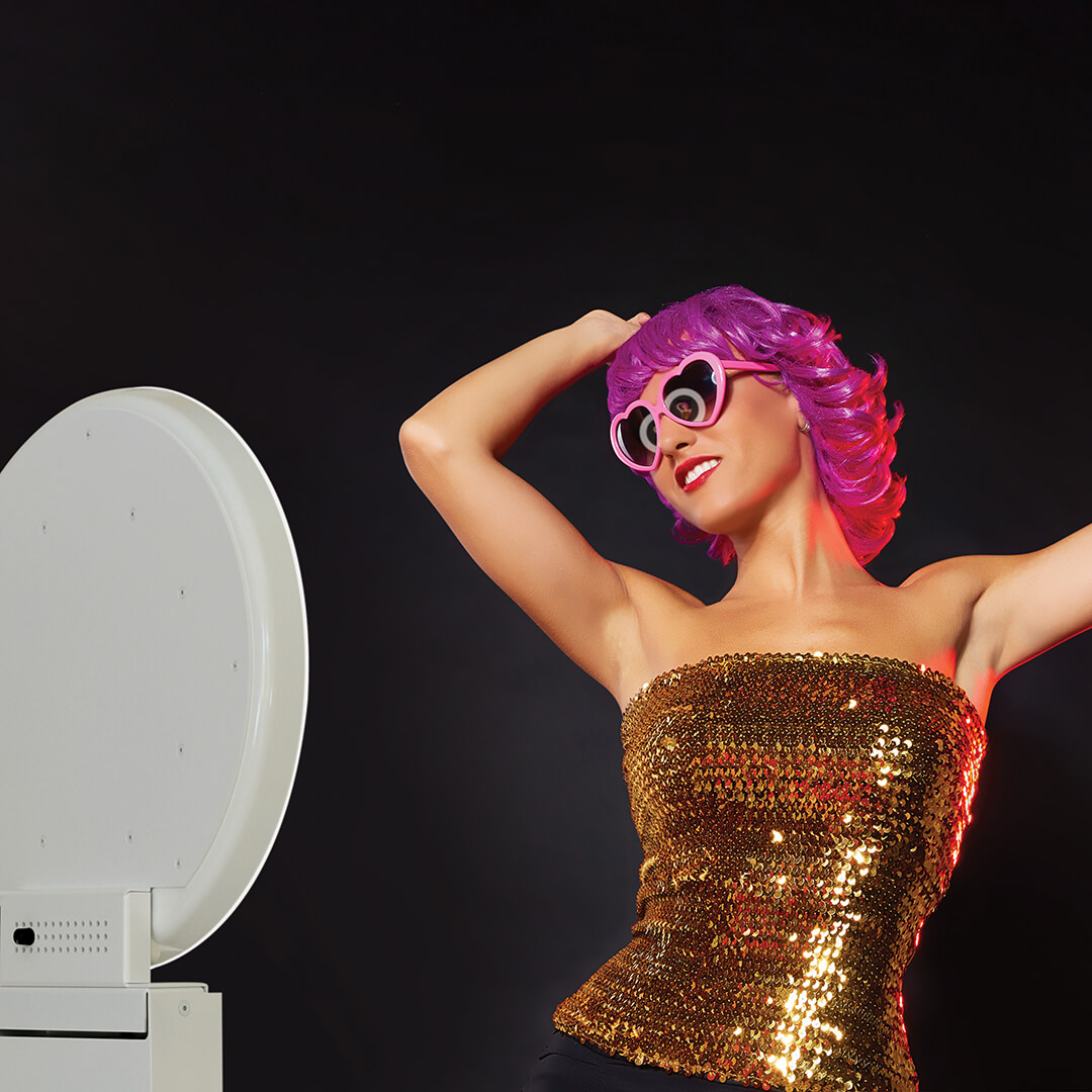 A woman with pink hair in heart shaped sunglasses and a gold top poses with her arms in the air in front of a white open air photo booth