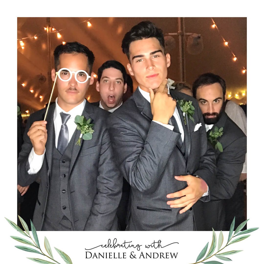 Four groomsmen pose in the photo booth while standing in a tent with string lights hanging overhead. One man is holding glasses on a stick as a prop.