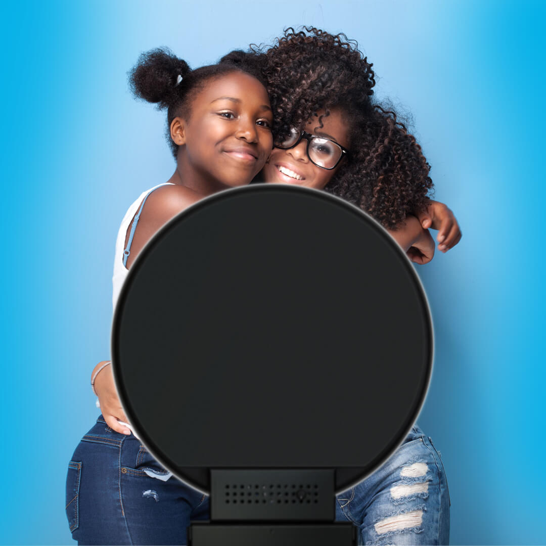 Two friends hug while posing against a blue backdrop in front of a photo booth. 